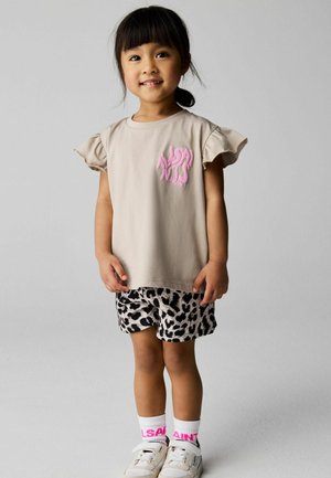 Młoda dziewczyna z czarnymi włosami związanymi w kucyk, ubrana w beżową koszulkę z falbanami na rękawach, czarno-białe szorty w zwierzęcy wzór, białe skarpetki i trampki.