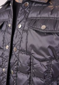 Veste noire rembourrée avec un design matelassé, dotée de boutons-pression argentés et de deux poches avant. Texture lisse avec un col et une fermeture éclair.
