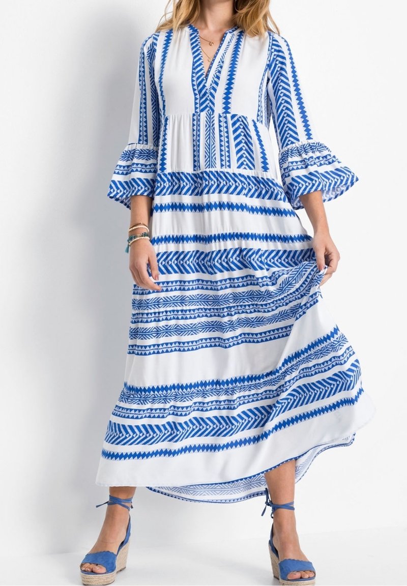 Robe maxi blanche et bleue avec des motifs géométriques. Dotée de manches évasées et d'un décolleté en V. Portée avec des sandales espadrilles bleues.