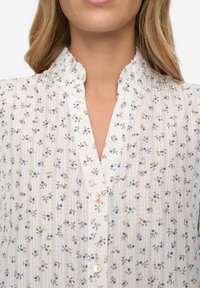 Witte blouse met bloemenprint met kleine blauwe en beige bloemen, verticale strepen, een gerimpelde kraag en een knoopsluiting met parelachtige knopen.