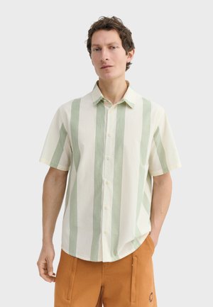 Hombre con camisa blanca de manga corta con botones y rayas verticales verdes, y pantalones marrones, de pie frente a un fondo claro y liso.