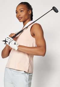 Růžový bezrukávový sportovní top s bočními výřezy, spárovaný se světle modrými kalhotami. Golfista drží černou a stříbrnou hůl v bílé rukavici.