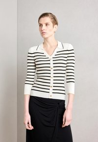 Gilet côtelé avec des rayures horizontales noires et blanches, décolleté en V, manches trois-quarts, fermeture à boutons dorés et poignets avec accents dorés.