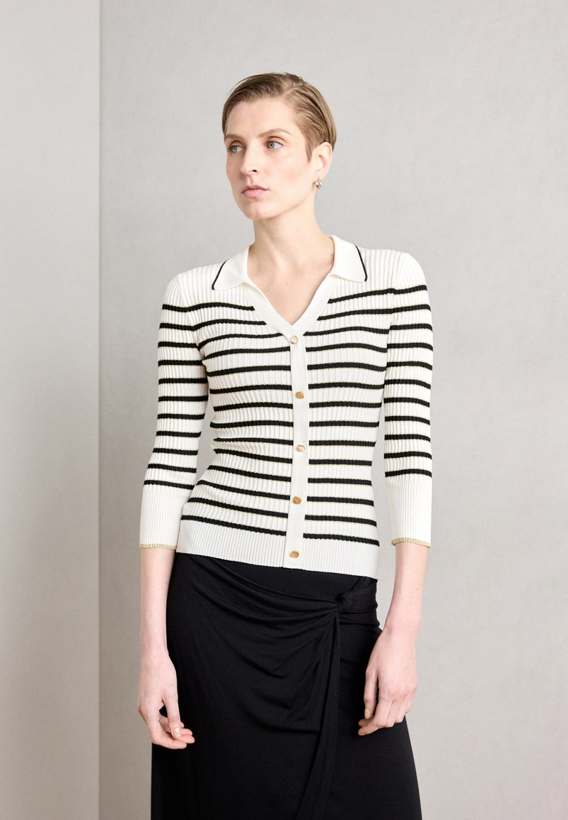 Gilet côtelé avec des rayures horizontales noires et blanches, décolleté en V, manches trois-quarts, fermeture à boutons dorés et poignets avec accents dorés.