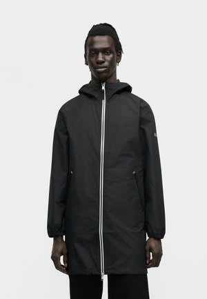 Jeune homme portant un imperméable noir à capuche avec une fermeture éclair frontale et des poches latérales zippées, debout devant un fond clair uni.