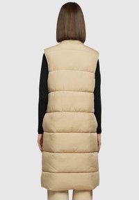 Beige puffervest met een lange ontwerp, voorzien van horizontaal gestikte secties, mouwloos snit en een gladde, matte afwerking.