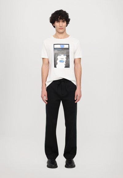 White cotton t-shirt featuring a graphic print of a woman's face in black and blue. Paired with black trousers that have green side stripes.
