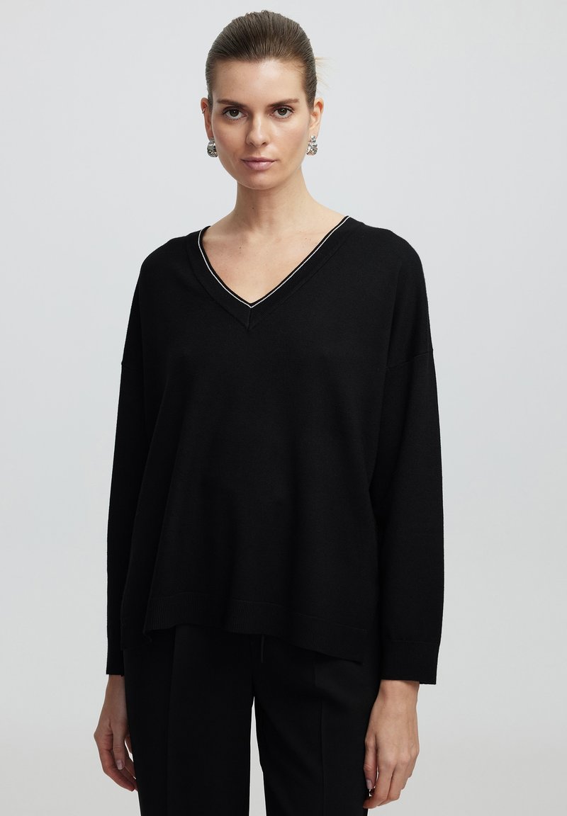 Pull en maille noir avec un décolleté en V et des coutures blanches contrastantes. Doté de manches longues et d'une coupe décontractée, associé à un pantalon noir.