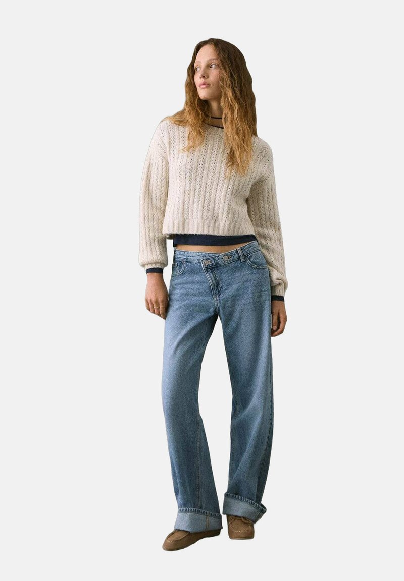 Maglione bianco a collo alto, lavorato a trecce, con maniche lunghe, abbinato a jeans blu chiaro, larghi, e scarpe marroni senza lacci.