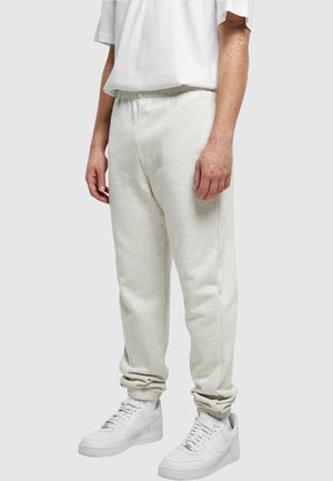 Personne portant un pantalon de survêtement gris clair, des baskets blanches et un t-shirt blanc à manches courtes, debout devant un fond uni.