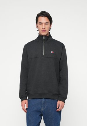 Meesmodell, kes kannab tumedat halli veerandilahti särk, millel on rindkerel väike Tommy Jeans logo, ja siniseid teksaseid, seistes valge tausta ees.