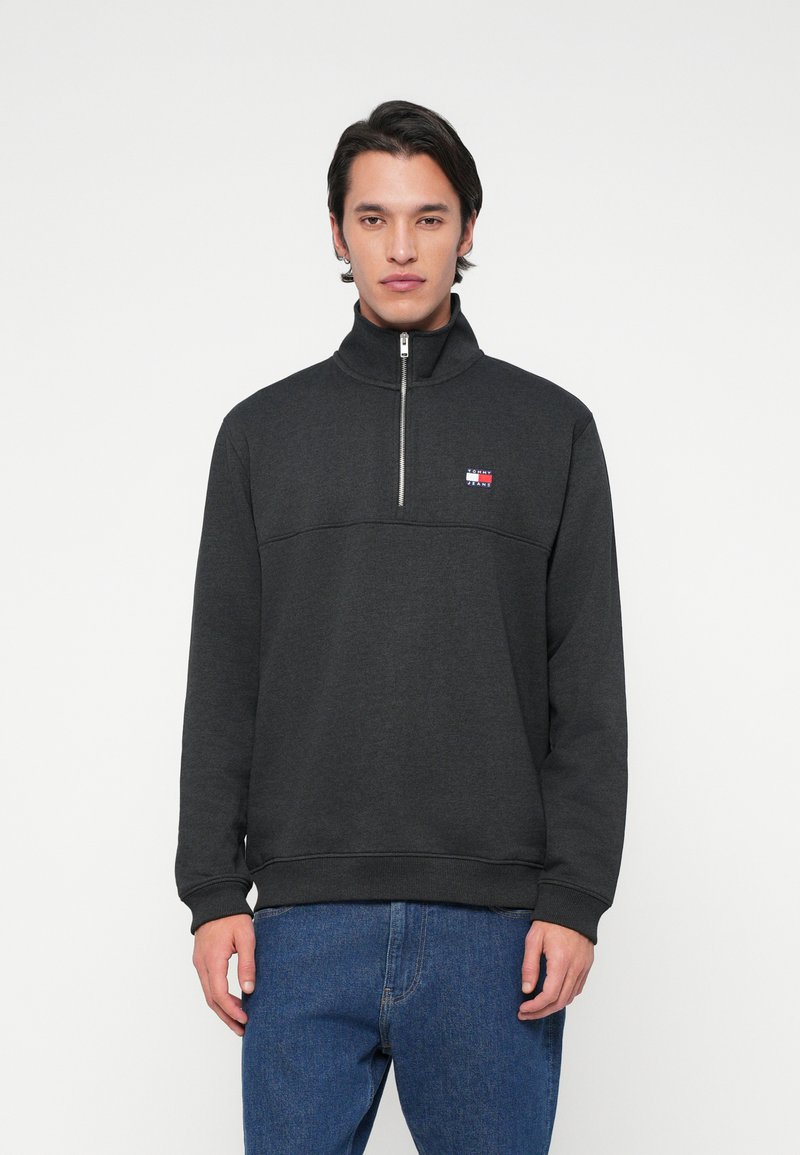 Άνδρας μοντέλο φοράει έναν σκούρο γκρι φούτερ με κοντό φερμουάρ και μικρό λογότυπο Tommy Jeans στο στήθος, συνδυασμένο με μπλε τζιν, στέκεται μπροστά σε λευκό φόντο.