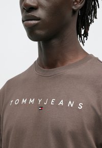 Bruin katoenen T-shirt met "TOMMYJEANS" geborduurd in wit en een klein logo-accent in rood, wit en blauw. Ontwerp met ronde hals.