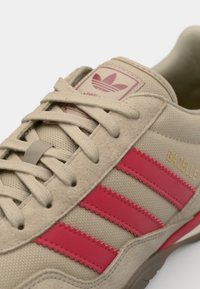 Χακί σουέτ αθλητικά παπούτσια με κόκκινες ελαστικές λωρίδες, υφή στο πάνω μέρος και τονικό κορδόνι. Διαθέτει ετικέτα με λογότυπο και το κείμενο "Gazelle" σε χρυσό χρώμα.