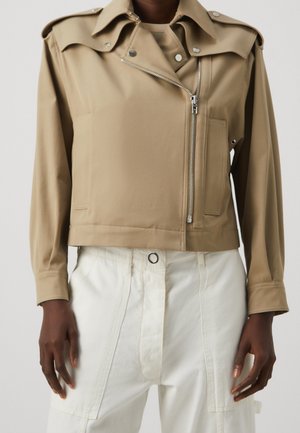 Personne portant une veste beige courte avec une fermeture éclair asymétrique et un pantalon blanc taille haute avec de grandes poches avant.