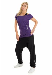Paarse t-shirt met korte mouwen en een ontspannen pasvorm, gecombineerd met losse zwarte harembroek. Het materiaal lijkt zacht. Wit logo detail op het shirt.