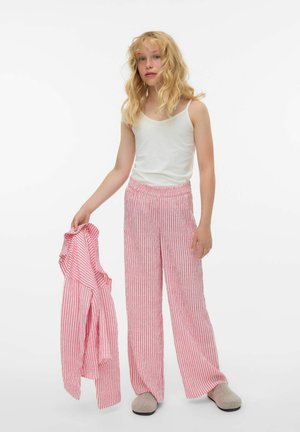 Hvit ermeløs topp, røde og hvite stripete vidbukser med elastisk midjebånd, holder matchende stripete skjorte. Grå tøfler, lyst hår.