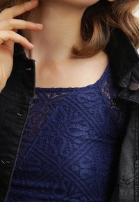 Donna che indossa un top di pizzo blu navy sotto una giacca di jeans nera, con una mano che sistema il colletto della giacca vicino al collo.