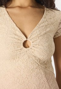 Robe en dentelle couleur nude avec un décolleté en V et un détail découpé circulaire avec une texture froncée au niveau de la poitrine et des manches courtes.