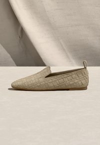 Μπεζ επίπεδο παπούτσι τύπου slip-on με υφή πλεγμένου δέρματος και μικρή ταμπελίτσα στο πίσω μέρος, τοποθετημένο πάνω σε ανοιχτόχρωμη επιφάνεια με φόντο μπεζ ύφασμα.