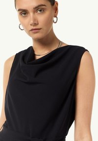 Czarny bezrękawnik z dekoltem w drapowania i gładkim materiałem. Podkreślony cienkimi metalowymi naszyjnikami i kolczykami w kształcie kółek.