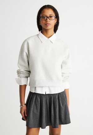 Nuori nainen, jolla on mustat silmälasit, vaaleanharmaa lyhyt collegepaita valkoisen kauluspuseron päällä ja tummanharmaa ohuilla raidoilla koristeltu laskostettu hame.