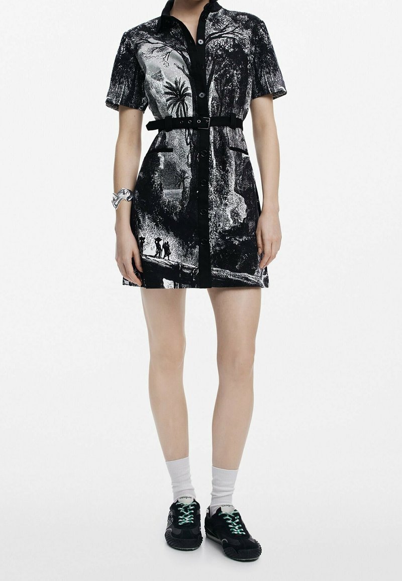 Robe noire et blanche à manches courtes avec fermeture à boutons sur le devant, imprimé nature, ceinture noire, chaussettes blanches, baskets noires et bracelet argenté au poignet gauche.