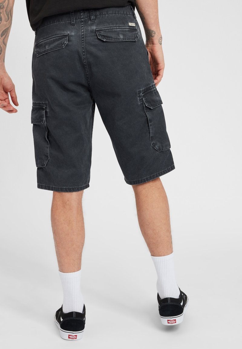 Shorts cargo noirs avec plusieurs poches, une coupe décontractée et une texture légèrement décolorée. Portés avec des chaussures noires et des chaussettes blanches.