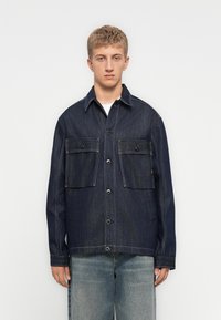 Jonge man in een donkerblauwe denimjack met grote klepzakken en lichtblauwe versleten spijkerbroek, staand tegen een plain witte achtergrond.