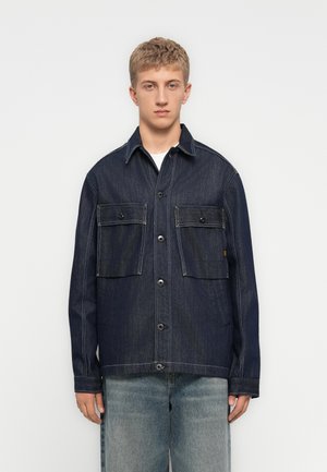 Jonge man in een donkerblauwe denimjack met grote klepzakken en lichtblauwe versleten spijkerbroek, staand tegen een plain witte achtergrond.