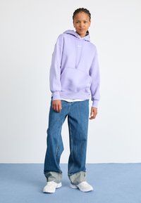 Um hoodie lavanda com um bolso frontal e cordão, combinado com jeans azuis largos. Ténis brancos completam o visual casual.