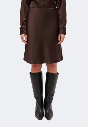 Femme portant une jupe en satin brun foncé et une chemise assortie, associées à des bottes en cuir noires montantes, debout devant un fond uni.