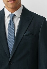 Námornícky textúrovaný sako s klopami, v kombinácii s bielou košeľou a modrou vzorovanou kravato. Kapsička v zodpovedajúcej látke.