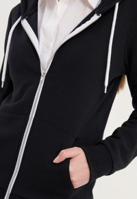 Personne portant un sweat à capuche noir zippé avec des cordons blancs et un t-shirt blanc en dessous, main reposant dans la poche du sweat.