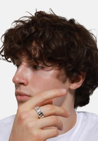 Anello in argento su una mano con tono di pelle chiaro, caratterizzato da una finitura liscia e lucida. Il modello ha i capelli ricci e scuri ed indossa una camicia bianca.