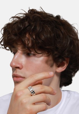 Silver chain ring on a hand with light skin tone, featuring a smooth, polished finish. Model has curly dark hair and is wearing a white shirt.