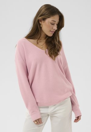 Žena s dlhými hnedými vlasmi, ktorá nosí zlaté kruhové náušnice, vrstvené náhrdelníky, voľný ružový sveter s výstrihom do V a biele nohavice, pozerá sa nadol.