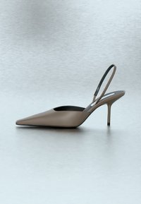 Γόβα slingback μυτερή σε χρώμα ταουπέ με λεπτό λουράκι στον αστράγαλο και λεπτό τακούνι στιλέτο σε ανοιχτό γκρι επιφάνεια.