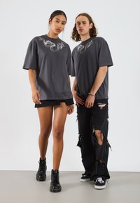 Deux individus portent des t-shirts oversized gris foncé avec un motif de serpent. La femme a une jupe noire superposée ; l'homme porte un jean noir déchiré et des baskets.