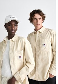 Camisas de pana beige con bolsillos frontales y logotipos; tejido texturizado; llevadas por dos modelos, uno con gorra y el otro con el cabello despeinado.