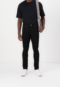Persona vestida con camiseta negra, jeans negros, zapatillas blancas y bolsa gris al hombro contra un fondo blanco.