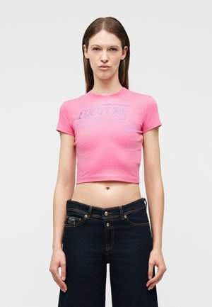 Naine seisab ühevärvilise tausta ees, kandes roosat lühikest T-särki siniste tekstidega ja tumedate kõrge vöökohaga teksapükse.