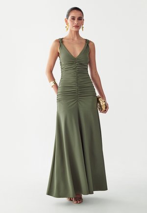 Vrouw draagt een geplooide olijfgroene maxi-jurk met geknoopte bandjes, gouden oorbellen, armband, en houdt een gouden clutch vast, staand op een witte achtergrond.