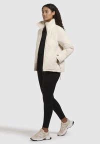 Chaqueta acolchada de color claro con un alto cuello, bolsillos laterales y cordones ajustables en el dobladillo, combinada con leggins negros y zapatillas deportivas.