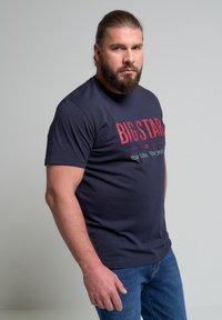 Camiseta de algodón azul marino con "BIG STAR" en impresión roja, de manga corta y cuello redondo. Lleva puesta con jeans de mezclilla azules.