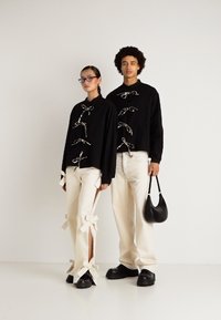 Du modeliai, vilkintys juodas ilgomis rankovėmis viršūnes su užsegimais ir kreminiais kelnais, iš kurių vienas su kaspinų akcentais, stovi prieš neutralų foną.