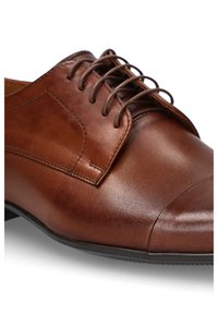 Brązowe skórzane buty formalne o gładkiej fakturze, zaokrąglonym nosku i sznurowaniu. Zawierają haftowany detal po bokach oraz czarną gumową podeszwę.