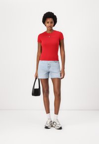 Rød kortermet tettsittende t-skjorte med logodetalj, kombinert med lys blå denimshorts. Modellen holder en liten svart håndveske. Sølvfargede joggesko er på føttene.