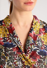Bluzka w kwiatowy wzór z dekoltem kopertowym, wykonana z lekkiego materiału. Kolory to czerwień, żółć, niebieski i zieleń na białym tle.