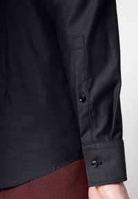 Czarny rękaw teksturowanej koszuli z dwoma czarnymi guzikami przy mankiecie. Tkanina ma subtelny wzór i gładkie wykończenie, uzupełnione zaokrąglonym dołem.